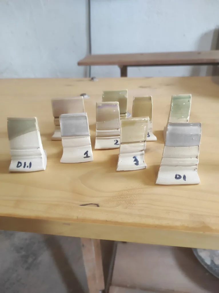 Fichas de prueba de esmaltes cerámicos en tonos verdes, grises y beige sobre mesa de madera